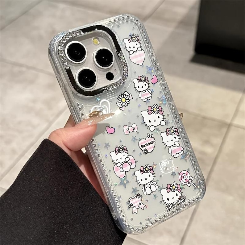 May include: Clear phone case with a sparkly, glittery design. The case features a pattern of Hello Kitty characters, pink hearts, bows, and stars. The edges are lined with small, clear rhinestones. The text "Good day!" and "Hello" are visible.