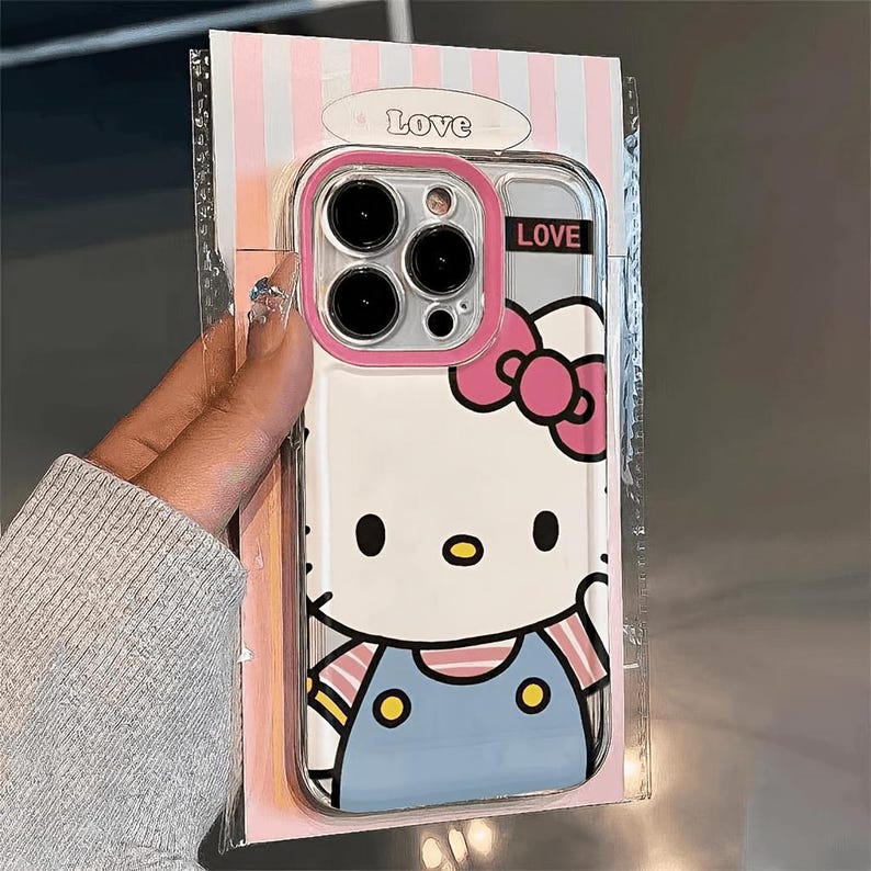 Pode incluir: Capa de telefone transparente com design Hello Kitty, la&ccedil;o rosa e macac&atilde;o azul. A capa tem uma borda rosa ao redor da c&acirc;mera e a palavra "LOVE" &eacute; impressa na capa. A capa &eacute; embalada em um suporte listrado rosa e branco.
