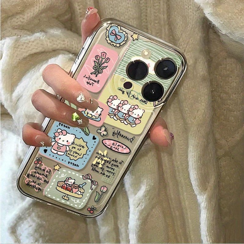 May include: Clear phone case with colorful stickers featuring cartoon characters, flowers, and text. The case has a clear border and a multi-lens camera cutout. The stickers are in shades of pink, blue, and yellow.