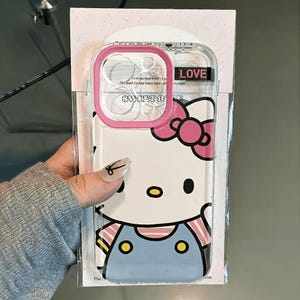 Pode incluir: Capa de telefone transparente com borda rosa, com um design Hello Kitty. A capa tem um fundo branco com uma imagem de desenho animado de Hello Kitty usando um la&ccedil;o rosa e macac&atilde;o azul. A palavra "LOVE" est&aacute; impressa na capa.
