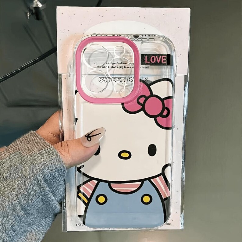 Pode incluir: Capa de telefone transparente com borda rosa, com um design Hello Kitty. A capa tem um fundo branco com uma imagem de desenho animado de Hello Kitty usando um la&ccedil;o rosa e macac&atilde;o azul. A palavra "LOVE" est&aacute; impressa na capa.