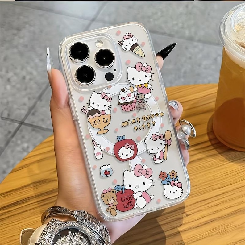 May include: Clear phone case with a colourful design featuring the character Hello Kitty, ice cream, cupcakes, and apples. The case has the words "Mint Green Kitty" and "Good Luck" printed on it. The case is held by a person.