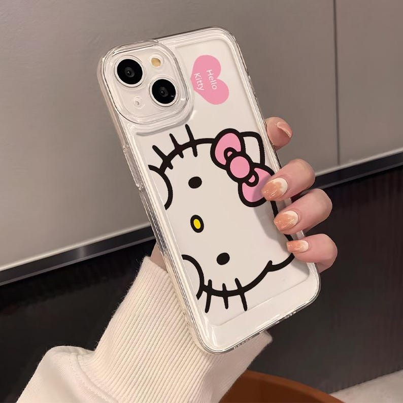 以下が含まれることがあります： ハローキティのデザインが施された透明な携帯電話ケース。ケースは白い背景に、ハローキティの顔の黒い輪郭、ピンクのリボン、ピンクのハートに「Hello Kitty」の文字が入っています。ケースには透明な縁があります。