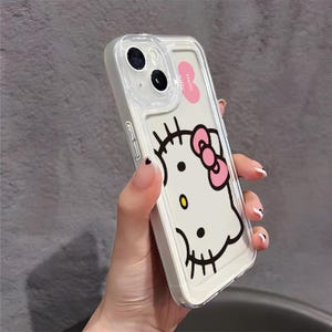 以下が含まれることがあります： ハローキティのデザインが施された透明な携帯電話ケース。ケースは、ピンクのリボン、黒いひげ、黄色の鼻が付いた白い背景です。ケースには透明な縁と「Hello Kitty」と書かれたピンクのラベルが付いています。