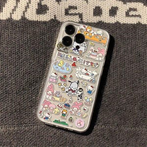 May include: Clear phone case decorated with colorful cartoon character stickers. The case features a variety of designs, including Hello Kitty, ducks, and other cute illustrations. The case has a clear back and black camera lenses.
