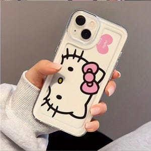 以下が含まれることがあります： 白い背景にハローキティのデザインが施された透明な携帯電話ケース。デザインには、ハローキティの顔の黒いアウトライン、ピンクのリボン、ピンクのハートの「Hello Kitty」という文字が含まれています。ケースには透明なボーダーがあります。