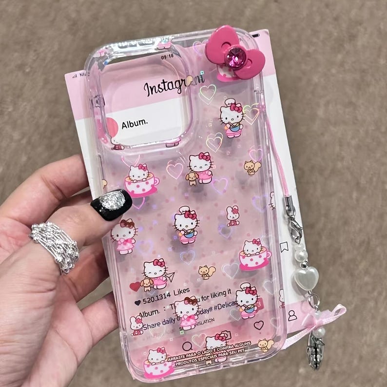 May include: Clear phone case with a pink bow and a Hello Kitty design. The case features illustrations of Hello Kitty, hearts, and teacups. The case has a pink strap with charms and the text "Instagrani" and "Album."