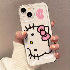以下が含まれることがあります： ハローキティのデザインが施された透明な携帯電話ケース。ケースは白い背景にピンクのリボンとハローキティのキャラクターが描かれています。「ハローキティ」の文字がピンクのハートにプリントされています。