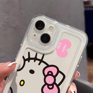 以下が含まれることがあります： ハローキティのデザインが施された透明な携帯電話ケース。ケースには「Hello Kitty」の文字が入ったピンク色のハートと、ピンクのリボンをつけたハローキティの漫画の画像があります。ケースは携帯電話のカメラを保護するように設計されています。