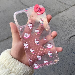 May include: Clear phone case featuring a pink bow and a pattern of Hello Kitty characters and pink polka dots. The case has a clear border and a pink bow with a gem in the center.