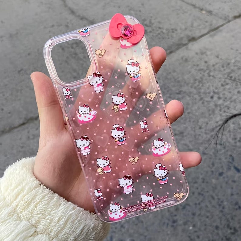May include: Clear phone case featuring a pink bow and a pattern of Hello Kitty characters and pink polka dots. The case has a clear border and a pink bow with a gem in the center.