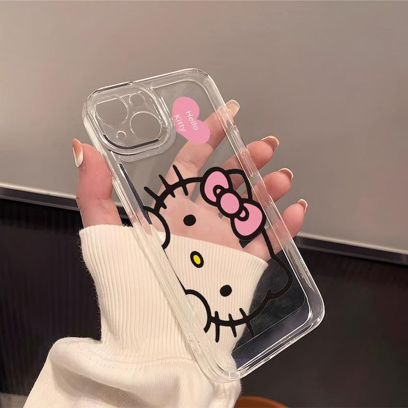 以下が含まれることがあります： ハローキティのデザインが施された透明な携帯電話ケース。ケースにはピンクのリボン、"Hello Kitty"の文字が入ったピンクのハート、黒いディテールがあります。このケースはスマートフォンに合うように設計されています。
