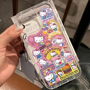 May include: Clear phone case decorated with Hello Kitty stickers in various poses and settings. The stickers feature pink, yellow, and white colors, with text like "HELLO KITTY" and "YEAH!". The case has a transparent design.