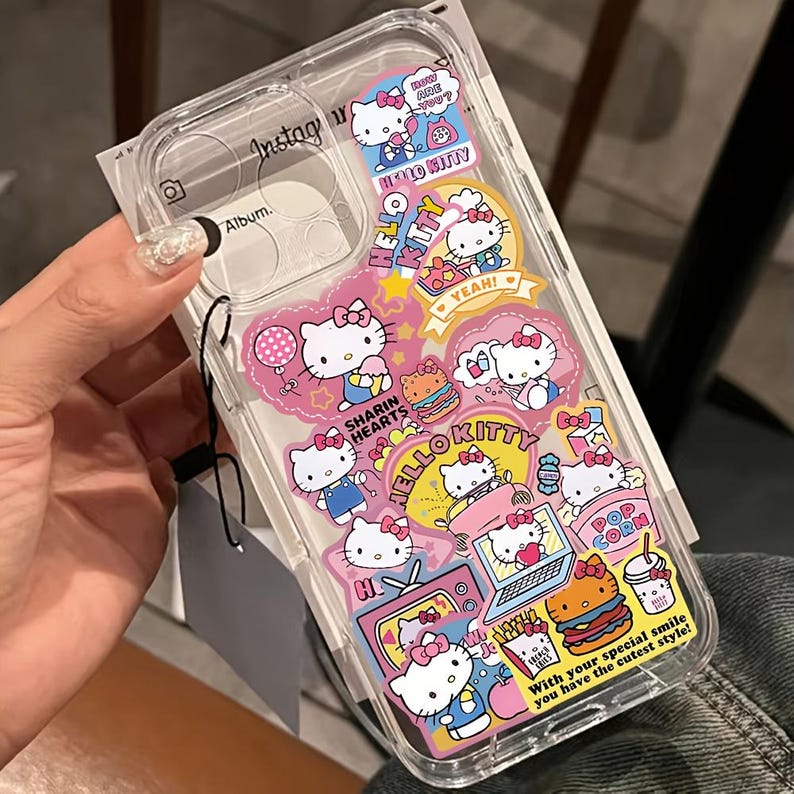 May include: Clear phone case decorated with Hello Kitty stickers in various poses and settings. The stickers feature pink, yellow, and white colors, with text like "HELLO KITTY" and "YEAH!". The case has a transparent design.