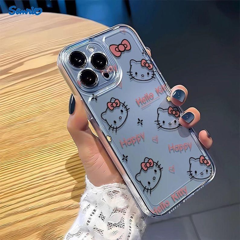 May include: Clear phone case featuring a repeating pattern of Hello Kitty faces with pink bows. The case also has the words "Happy" and "Hello Kitty" in pink. The case is held in a hand.