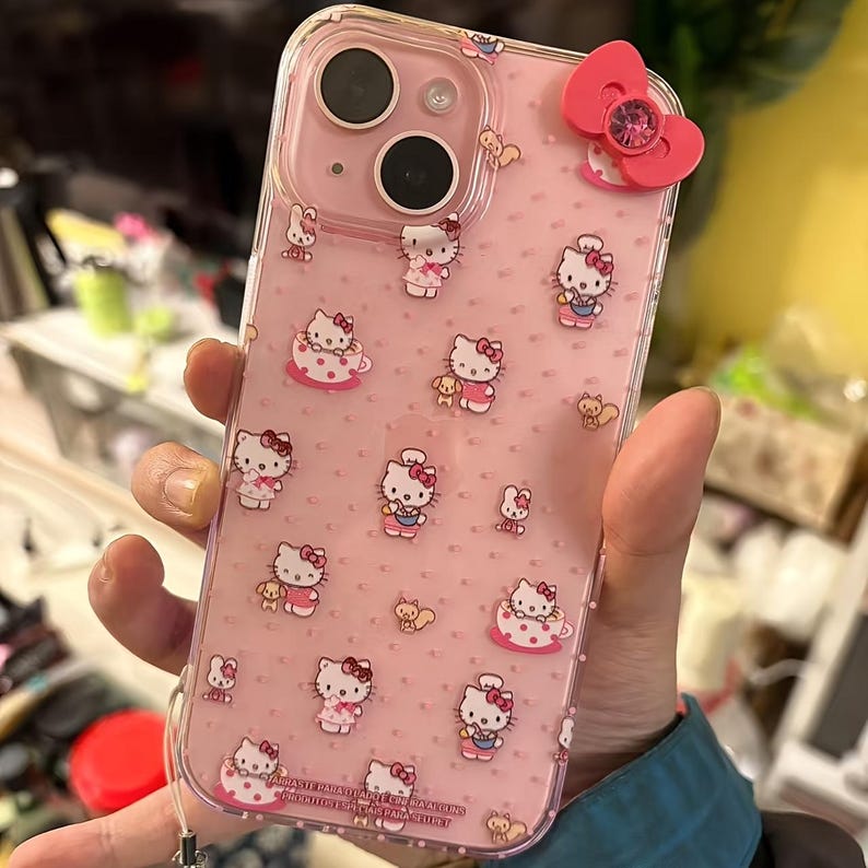 May include: Pink phone case with a clear border, featuring a repeating pattern of Hello Kitty characters in various poses. A pink bow with a crystal center adorns the top. The case is designed for a smartphone.