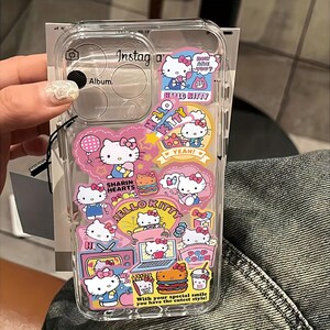 May include: Clear phone case decorated with Hello Kitty stickers. The stickers feature Hello Kitty characters with various accessories and text like "YEAH!" and "How are you?". The case also has the text "With your special smile you have the cutest style!"