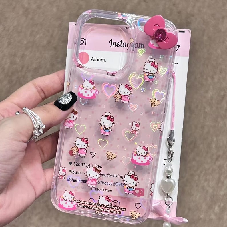 May include: Clear phone case featuring Hello Kitty designs, pink hearts, and a pink bow. The case has a pink strap with a pearl and heart charm. The case has the text "Insta&ccedil;yam" and "Album."