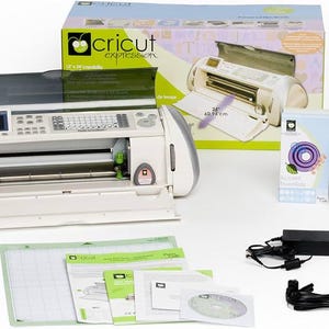 May include: A white Cricut Expression electronic cutting machine with a 12x24 inch cutting area. The machine is open and shows the cutting blade. The machine is surrounded by accessories, including a cutting mat, power adapter, and instruction manuals. The box for the machine is also visible in the background.