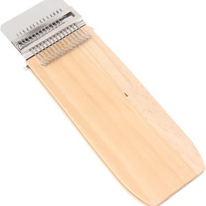 May include: A wooden kalimba instrument with a light wood finish. It features a metal tine bridge and a series of metal tines for plucking. The instrument has a rectangular shape with rounded edges and a silver metal cover.