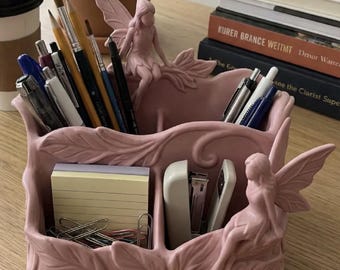 May include: A pink desk organizer with a fairy theme. The organizer has multiple compartments for pens, pencils, sticky notes, and paper clips. Two fairy figurines are perched on the sides. A stapler is also visible.