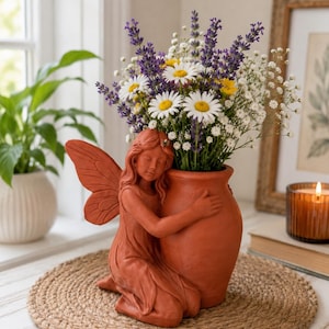 May include: A terracotta-colored ceramic planter shaped like a kneeling fairy with wings, embracing a rounded vase. The vase is filled with a bouquet of purple lavender, white baby's breath, and white daisies with yellow centers.