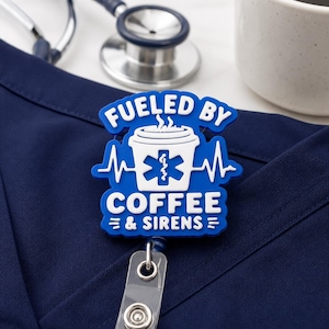 Puede incluir: Un portatarjetas azul con un gráfico de taza de café blanca, un símbolo médico de estrella de la vida, una línea de latido y el texto FUELED BY COFFEE & SIRENS. El portatarjetas está unido a una correa de plástico transparente y un broche metálico.