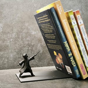 Può includere: Un fermalibri in metallo nero con una statuetta di mago che tiene una bacchetta, che sostiene una pila di libri colorati. I libri hanno titoli in polacco. Il fermalibri è su una superficie grigia.