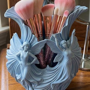 Puede incluir: Un soporte decorativo azul claro para brochas de maquillaje con dos figuras de ángeles esculpidas. El soporte contiene varios pinceles de maquillaje con cerdas rosas y blancas y mangos dorados. El soporte está sobre una superficie de madera.