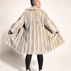 May include: A light grey fur coat with a wide collar and long sleeves. The coat is knee-length and has a flared silhouette. The person is wearing black leggings and black heels.