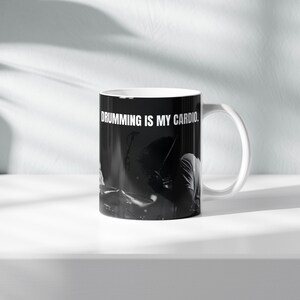 Könnte beinhalten: Ein weißer Keramikbecher mit einem schwarz-weißen Fotodruck eines Schlagzeugers an einem Schlagzeug. Der Text DRUMMING IS MY CARDIO ist in fetten, weißen Großbuchstaben im oberen Bereich der Außenseite des Bechers aufgedruckt.