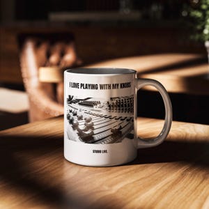 Puede incluir: Taza de cerámica blanca con una imagen en blanco y negro de una mesa de mezclas. La taza tiene el texto "I LOVE PLAYING WITH MY KNOBS" y "STUDIO LIVE". La taza está sobre una mesa de madera.