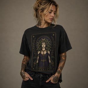 May include: A charcoal grey short-sleeve t-shirt featuring a rectangular graphic print of Medusa with green snakes for hair, wearing a dark purple dress. The design includes gold-toned accents, laurel branches, and the text MEDUSA at the bottom.