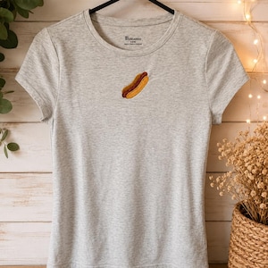 Peut inclure: Un t-shirt à manches courtes gris chiné clair avec un petit motif brodé de hot-dog centré sur la poitrine. L'étiquette intérieure du col affiche le texte No Boundaries et la taille S (3-5). Le t-shirt est présenté sur un cintre noir.
