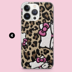 Puede incluir: Una funda para smartphone con estampado de leopardo en tonos beige y marr&oacute;n, decorada con personajes de gatos blancos bordados que llevan lazos de color rosa brillante. La funda tiene un recorte para una c&aacute;mara triple y el n&uacute;mero cuatro.