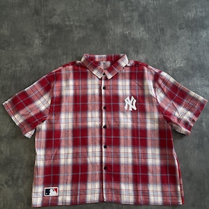 Könnte beinhalten: Rot-weiß kariertes Hemd mit kurzen Ärmeln. Es zeigt das Logo der New York Yankees auf der Brust und ein Logo der Major League Baseball am Saum. Das Hemd hat einen klassischen Kragen und eine Knopfleiste.
