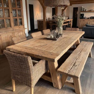 Könnte beinhalten: Ein rustikales Esszimmer-Set mit einem großen, verwitterten Holztisch, einer passenden Bank und Korbstühlen. Der Tisch hat eine natürliche Holzoberfläche, die Stühle haben hellfarbene Kissen. Das Set befindet sich in einem hellen, offenen Essbereich.