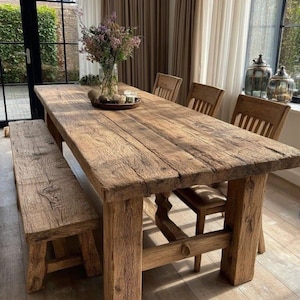 Op de afbeelding: Een rustieke houten eettafel met bijpassende bank en stoelen. De tafel is gemaakt van gerecycled hout en heeft een natuurlijke, verweerde afwerking. De stoelen hebben houten frames en leren zittingen. Een vaas met bloemen staat op de tafel.