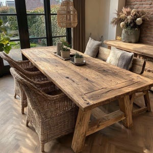 Könnte beinhalten: Ein großer, rustikaler Esstisch aus Holz mit natürlicher Oberfläche. Der Tisch ist von geflochtenen Korbstühlen und einer Bank umgeben. Dekorative Pflanzen und Kissen schmücken den Tisch. Eine gewebte Pendelleuchte hängt darüber und schafft einen warmen Essbereich.