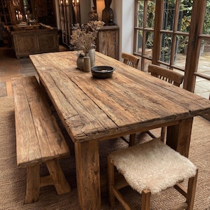 May include: A rectangular wooden dining table with a weathered, textured surface, accompanied by a matching wooden bench and a wooden chair featuring a white sheepskin seat cover. A dark bowl and two small vases with dried flowers sit on the table.
