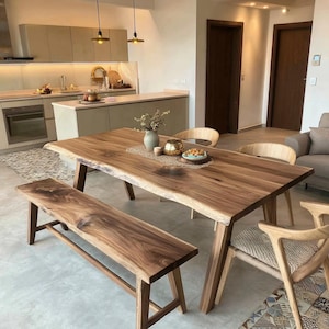 May include: A live edge walnut wood dining table with a matching bench and three light-coloured wooden chairs with woven seats. The table features a natural wood grain pattern and sits on angled legs, with a decorative runner and vase on top.