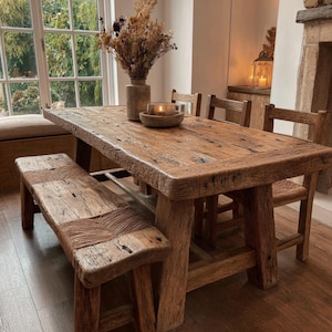 May include: A reclaimed wood dining table with a matching bench and chairs. The table and bench have a distressed finish, and the chairs have woven seats. A vase with dried flowers and a candle sit on the table. The room has a warm, inviting atmosphere.
