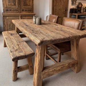 Könnte beinhalten: Ein rustikaler Esstisch aus Holz mit passender Bank und zwei Lederstühlen. Tisch und Bank bestehen aus Altholz und zeigen natürliche Maserung und Knoten. Zwei kleine Gläser stehen auf dem Tisch. Die Möbel haben eine warme, einladende Ästhetik.