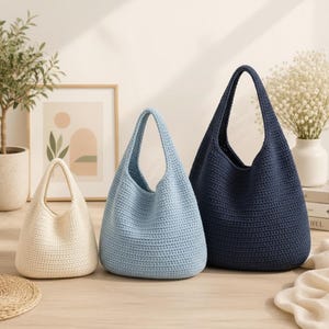 May include: Three crochet tote bags in cream, light blue, and navy blue. The bags have a rounded shape with a single handle. The bags are displayed on a light wood surface.