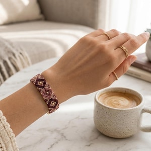 May include: A woven bracelet featuring a geometric diamond pattern in shades of burgundy, dusty rose, and cream, accented with small gold-toned beads at the center of each diamond shape, worn alongside three thin gold-toned metal rings on the fingers.