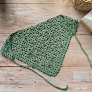 May include: A sage green crochet bag with a floral pattern. The bag has a drawstring closure and is made of a soft, textured yarn. The bag is lying on a wooden surface.