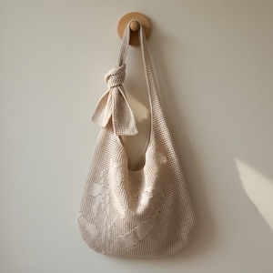 Puede incluir: Un bolso de hombro crema, de tejido abierto, con una correa anudada y detalles decorativos. El bolso está suspendido de una clavija de madera en una pared de tonos neutros, mostrando su textura y diseño. El color suave y el tejido aireado sugieren un accesorio informal.