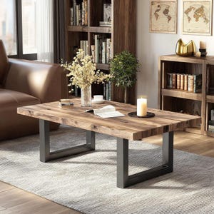 Puede incluir: Una mesa de centro rectangular de madera con un acabado de vetas naturales y patas de metal gris oscuro. La mesa está en un salón con un sofá de cuero marrón, estanterías y objetos decorativos.