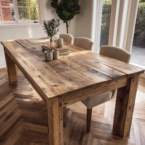 May include: A large, rectangular wooden dining table with a rustic, weathered appearance. The table is made of reclaimed wood and has a natural finish. Two beige upholstered chairs are partially visible. The table is approximately 183 cm long.