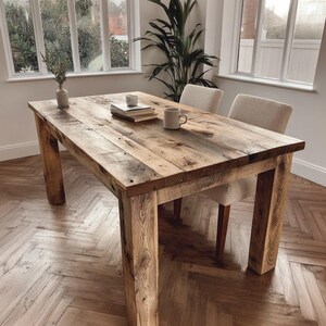 May include: A rustic wooden dining table with a natural finish. The table has a rectangular top and chunky legs. A vase, books, a mug, and a small candle holder are on the table. Two upholstered chairs are positioned behind the table.